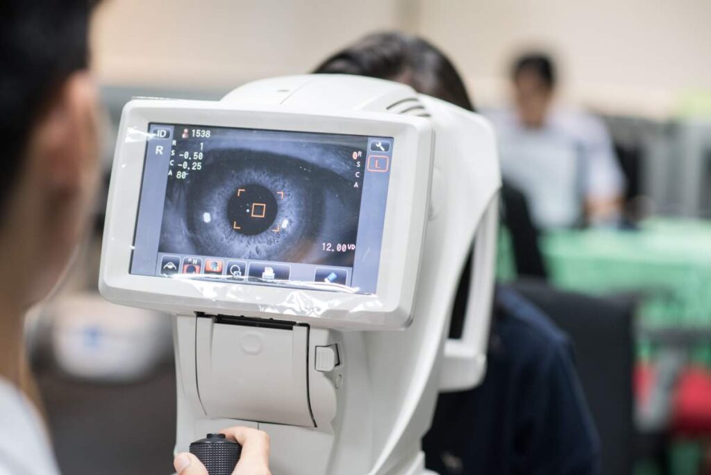 An optometrist examines the eyes of an adult patient using specialized equipment in a bright exam room.